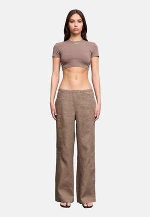 Donna in piedi di fronte, che indossa una t-shirt corta color taupe, pantaloni larghi marroni, sandali neri e una collana su uno sfondo semplice.