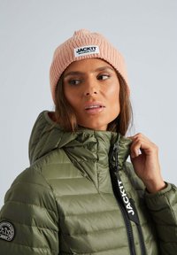 Roze gebreide beanie met ribbeltextuur en logo patch, gecombineerd met een gewatteerde olijfgroene jas met rits en capuchon.