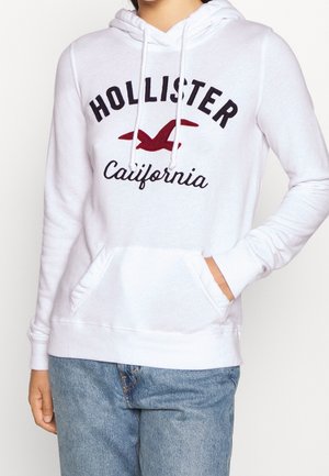 Biała bluza z kapturem z napisem „Hollister California” i czerwonym logo mewy, noszona przez osobę z ręką w kieszeni i niebieskimi dżinsami.