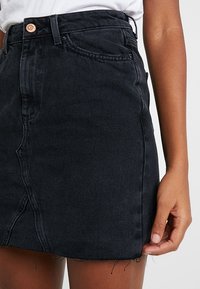 Mini-jupe en denim noir avec une taille haute, des coutures frontales et un ourlet effiloché. Dotée d'un bouton en cuivre et d'un design de poche traditionnel.