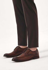 Scarpe da uomo eleganti in pelle marrone con texture liscia, design con lacci e dettagli di cuciture sottili. Abbinate a pantaloni sartoriali scuri.