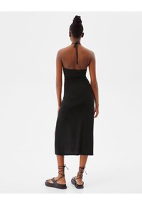 Svart, figurnära midiklänning med halterneck och knytning bak, med en slät textur och ankellångt snitt. Bärs med platta sandaler.