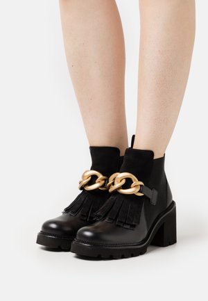 Des bottines noires présentent un talon épais, des accents en daim, des détails frangés et une chaîne dorée bien visible à l'avant. Matériau en cuir lisse.