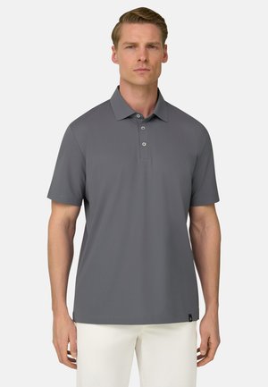 Mężczyzna ubrany w szarą koszulkę polo z krótkim rękawem i trzema guzikami oraz białe spodnie, stojący na jednolitym jasnym tle.