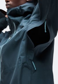 Gros plan d'une personne portant une veste bleu foncé avec un rabat de ventilation zippé sous le bras ouvert et des tirettes de fermeture éclair bleu clair.