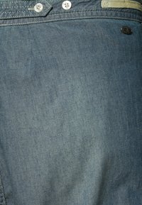 Tessuto di denim azzurro chiaro con una texture liscia; presenta due bottoni metallici e un'etichetta con logo. Il materiale ha un leggero lucido.