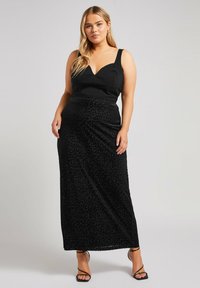 Schwarzes ärmelloses Oberteil mit tiefem V-Ausschnitt und einem langen, fließenden schwarzen Rock mit subtilen Leopardenmuster. Dazu werden dünne schwarze High Heels getragen.