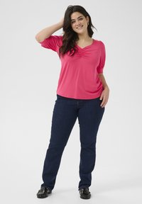 Haut rose à manches courtes avec un col froncé, associé à un jean bleu foncé et des chaussures noires à enfiler. Le modèle se tient de manière décontractée, une main sur la tête.