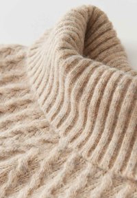 Beiger Strickpullover mit hohem Rippenkragen, der ein strukturiertes Muster aufweist und aus weichem, flauschigem Material gefertigt ist.