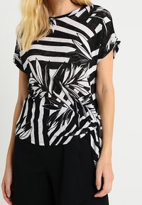 Czarny i biały top w paski z kwiatowym wzorem, krótkie rękawy, okrągły dekolt oraz węzeł w talii. Miękka tekstura materiału.