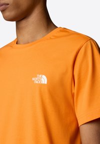 Orange bomulds T-shirt med rund hals og korte ærmer, med et hvidt logo af "The North Face" på venstre bryst.