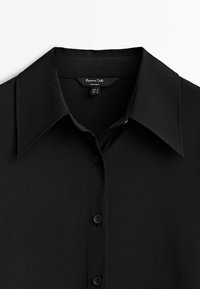 Chemise noire boutonnée en tissu lisse, avec un col pointu, six boutons sur le devant et une légère brillance.