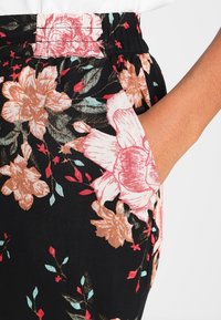 Handen i fickan på svarta tygbyxor med rosa, orange och grönt blommönster samt elastisk midja.