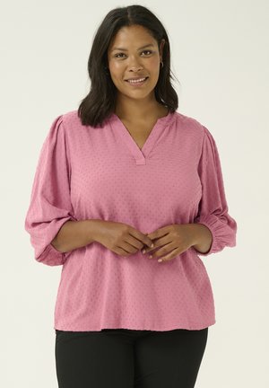 Top blouson rosa, tessuto testurizzato con scollatura a V e maniche arricciate; indossato con pantaloni neri. Il tessuto presenta una sottile fantasia a pois.