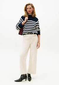 Frau mit einem navy- und weiß gestreiften Pullover, cremefarbenen weit geschnittenen Hosen, schwarzen spitzen Stiefeln, die eine burgunderfarbene Handtasche über der Schulter trägt.
