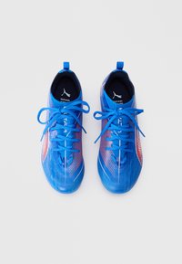 Par av blå Puma fotbollsskor med orange detaljer, blå snören knutna och Puma-logotyp synlig inuti hälområdet.