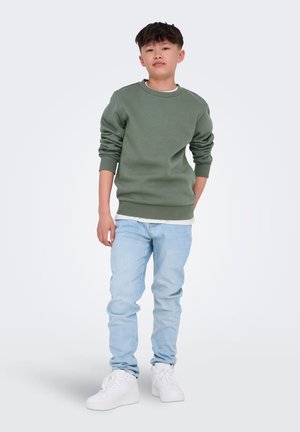 Felpa verde con polsini a coste, indossata sopra una camicia bianca, abbinata a jeans azzurri chiari e scarpe da ginnastica bianche. Posizione in piedi su uno sfondo chiaro.