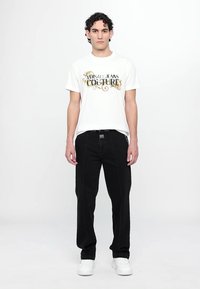 Balta medvilninė marškinėliai su užrašu "VERSACE JEANS COUTURE" spausdintu auksine ir juoda spalva. Juodos kelnės su baltomis siūlėmis. Modelis dėvi baltus sportbačius.