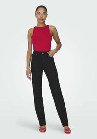 Röd ärmlös topp i kombination med höga svarta jeans. Modellen har på sig röda sandaler. Slät textur, åtsittande design och enfärgad mönster.