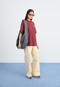T-shirt oversize bordeaux, pantalon large crème, sac à épaule gris décontracté, et baskets marron. Le mannequin porte des lunettes de soleil carrées.