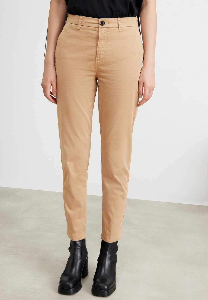 Personne portant un pantalon beige à jambes droites et des bottines noires à talons, debout sur un sol clair contre un mur blanc.