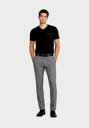 Bărbat în picioare cu mâinile în buzunare, purtând cămașă neagră cu decolteu în V, pantaloni gri eleganți, curea neagră și pantofi negri eleganți, pe un fundal simplu.