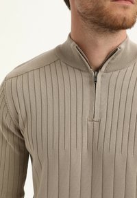Suéter acanalado en un beige claro con un cuello de media cremallera. La textura es suave, con rayas verticales y un diseño ajustado.