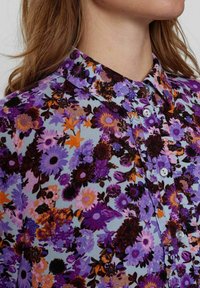 Bluză florală cu un fundal bleu deschis, decorată cu flori violete, portocalii și negre. Guler clasic și închidere cu nasturi, cu nasturi strălucitori.