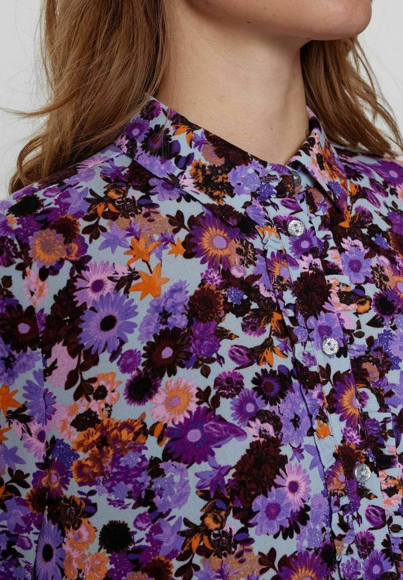 Blumenbluse mit hellblauem Hintergrund und lila, orangefarbenen sowie schwarzen Blumen. Klassischer Kragen und Knopfleiste mit glänzenden Knöpfen.