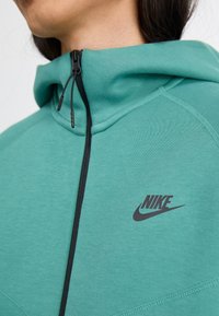 Mint green Nike hoodie with a black zipper, featuring a stitched design and logo on the chest. Soft fabric and a high collar design.