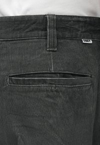 Tamsiai pilki keteriniai kelnių turi galinį kišenę su sklende, matomą prekinį ženklą ant juosmens ir tekstūruotą vertikalią raibą visame paviršiuje.