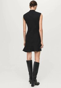 Black sleeveless dress with a fitted bodice and ruffled hem. Smooth fabric and a high neckline, paired with tall black leather boots.