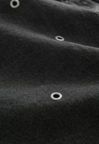 Tissu noir à surface texturée, orné d'œillets argentés. Le matériau semble durable et lourd, soulignant un design structuré.