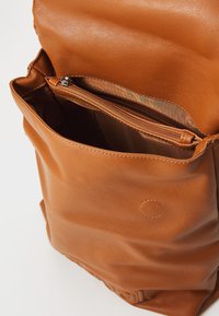 Intérieur d'un sac à dos en cuir marron avec une poche zippée. Texture douce et finition lisse. Détails de couture visibles en haut et logo.