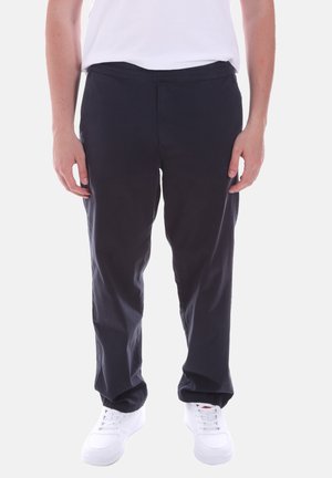 Persona che indossa pantaloni casual neri, sneaker bianche e una maglietta bianca, in piedi contro uno sfondo chiaro e semplice.