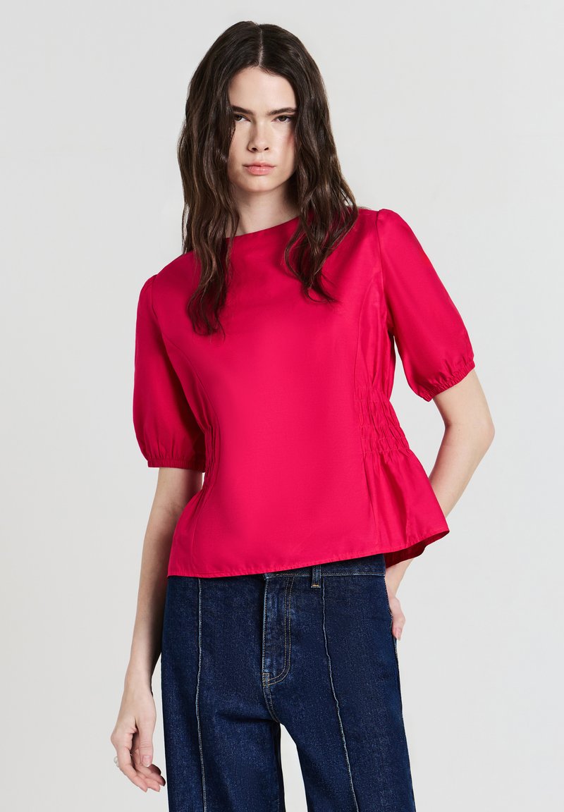 Donna con lunghi capelli scuri che indossa una blusa corta rosa brillante e jeans blu scuro, in piedi contro uno sfondo chiaro e uniforme.