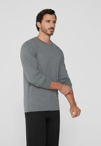 Mann mit dunklem Haar und Bart, der ein graues Langarmshirt und schwarze Hose trägt und seinen linken Ärmel vor einem weißen Hintergrund anpasst.