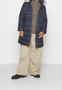 Marineblauer Steppmantel mit einem pelzgefütterten Kapuzen, kombiniert mit einem braunen Pullover und beigefarbenen, weit geschnittenen Hosen; strukturierte Materialien und kontrastierende Farben.