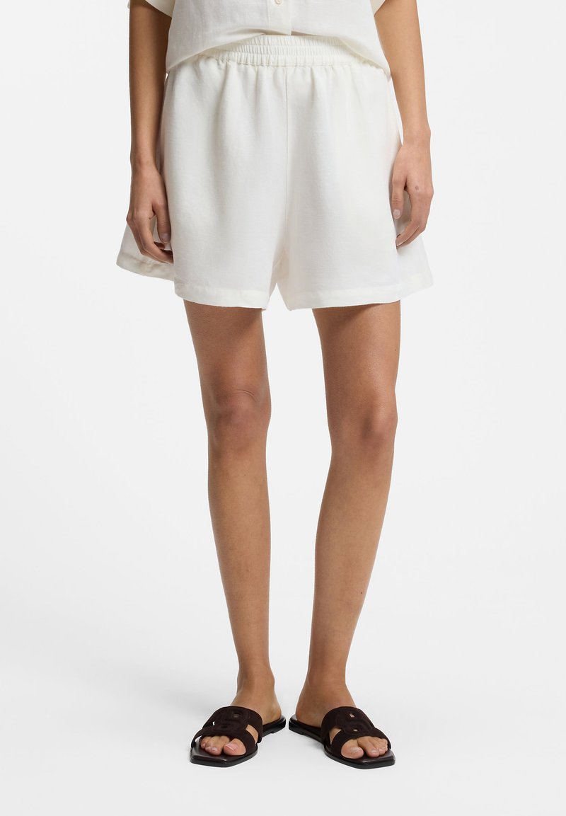 Γυναίκα που φοράει άνετα λευκά σορτς με ελαστική μέση και μαύρα φλατ σανδάλια τύπου slide σε λευκό φόντο.