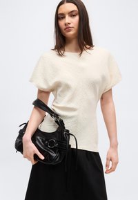 Γυναίκα με μακριά σκούρα μαλλιά, που φοράει μια υφασμάτινη κοντομάνικη μπλούζα εκρού και μαύρη φούστα, κρατώντας μια μαύρη δερμάτινη τσάντα στο αριστερό της χέρι.