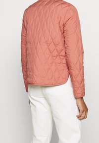 Giacca trapuntata in rosa antico, con un design a scatola e una texture liscia. Presenta un collo rotondo e maniche lunghe. Accompagnata da pantaloni bianchi.