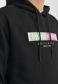 Zwarte hoodie met een grafisch ontwerp aan de voorkant met "JACK & JONES" in wit, paars en groen, met een trekkoord en een gestructureerde stof.