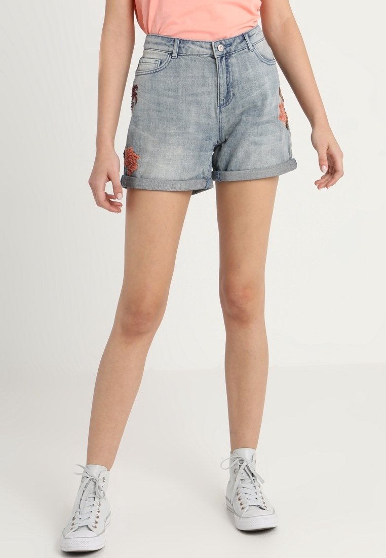 Pantalones cortos de mezclilla azul claro con dobladillos enrollados, con bordado floral rojo en los lados. Usados con zapatillas altas blancas.