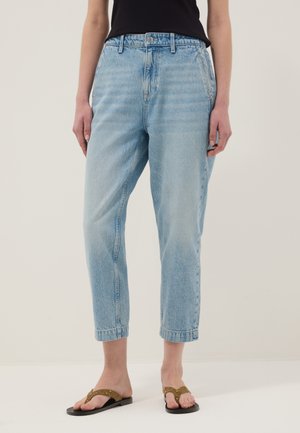 Persona que lleva vaqueros cortos de tiro alto de color azul claro, con una camiseta negra y sandalias marrones tipo chancla, de pie frente a un fondo liso.