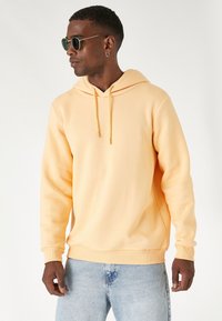 Kapturowa bluza w pastelowym pomarańczowym kolorze, wykonana z mieszanki bawełny. Posiada sznurki, ściągacze przy rękawach oraz luźny krój. Dżinsy w jasnym odcieniu.