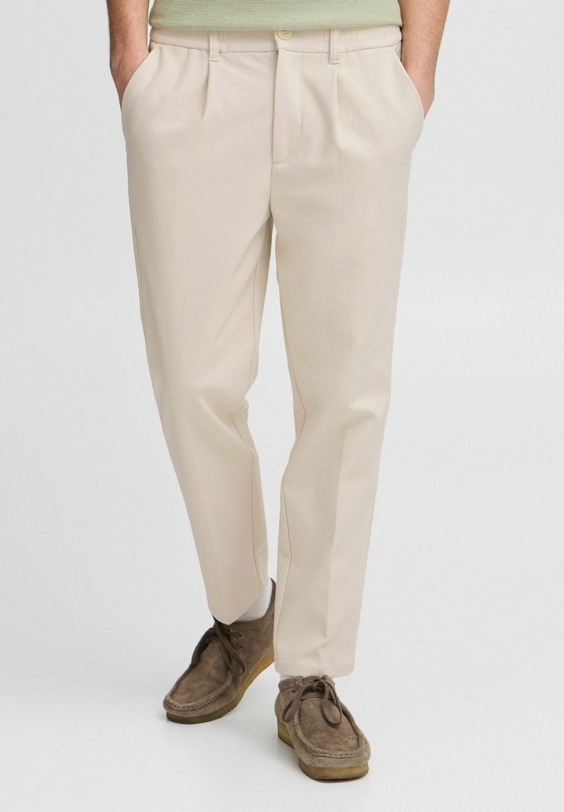 Man wearing beige pleated trousers and brown suede lace-up shoes with white socks, standing with hands in pockets.