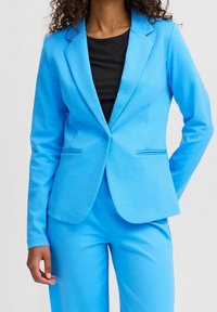 Kvinna iklädd en ljusblå skräddarsydd kavaj och matchande byxor tillsammans med en svart ribbad topp mot en enkel ljus bakgrund.