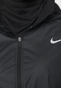 Czarna kurtka zapinana na zamek o gładkiej fakturze, z wysokim kołnierzem i odblaskowym białym logo na piersi. Wewnętrzna warstwa jest czarna.