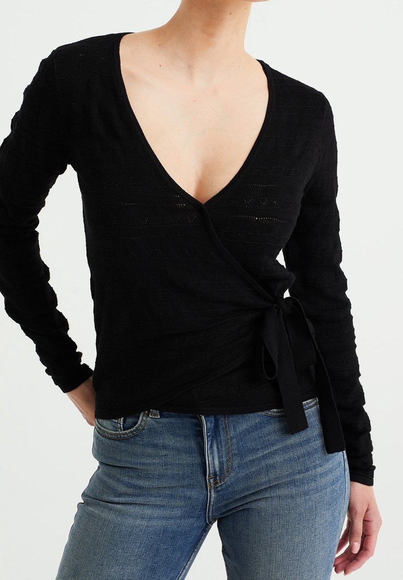 Haut en tricot noir avec un décolleté en V profond, des manches longues et un lien à la taille. Présente un motif texturé sur une surface lisse.