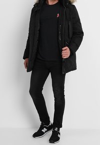 Black puffer jacket with fur-lined hood, zip and snap closures, side pockets, worn over a black shirt and paired with black jeans and sneakers.